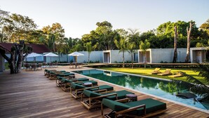 Zona de piscina con sillones, una piscina y un jardín con árboles y un edificio al fondo.