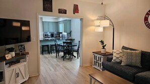 Un salon moderne avec un canapé foncé, une table basse et un téléviseur. On aperçoit un coin repas avec une table et des chaises à travers une porte ouverte.