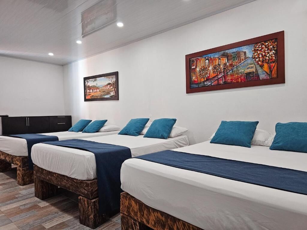 Habitación de hotel con tres camas individuales, cada una con cabecera azul y ropa de cama blanca. Hay dos cuadros en las paredes, uno que representa un paisaje y otro con una escena abstracta y colorida.