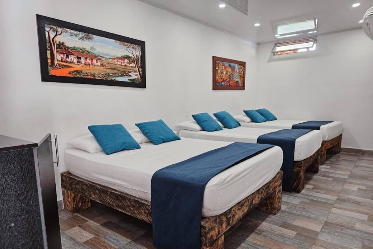 A hotel room with three beds, each with a blue blanket and pillow. There are two paintings on the wall, a window with a view, and a ceiling fan.