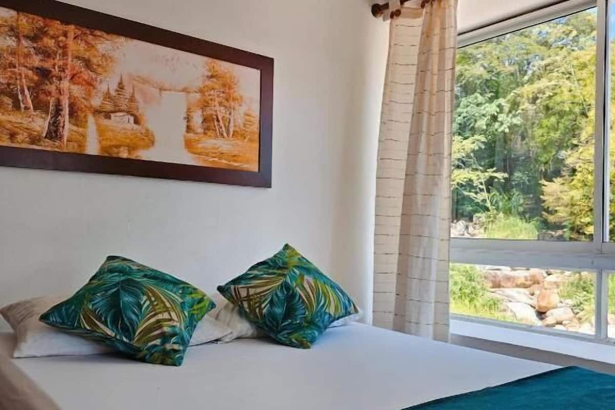 A bedroom with a bed, two pillows, a painting, and a window with a view of trees.