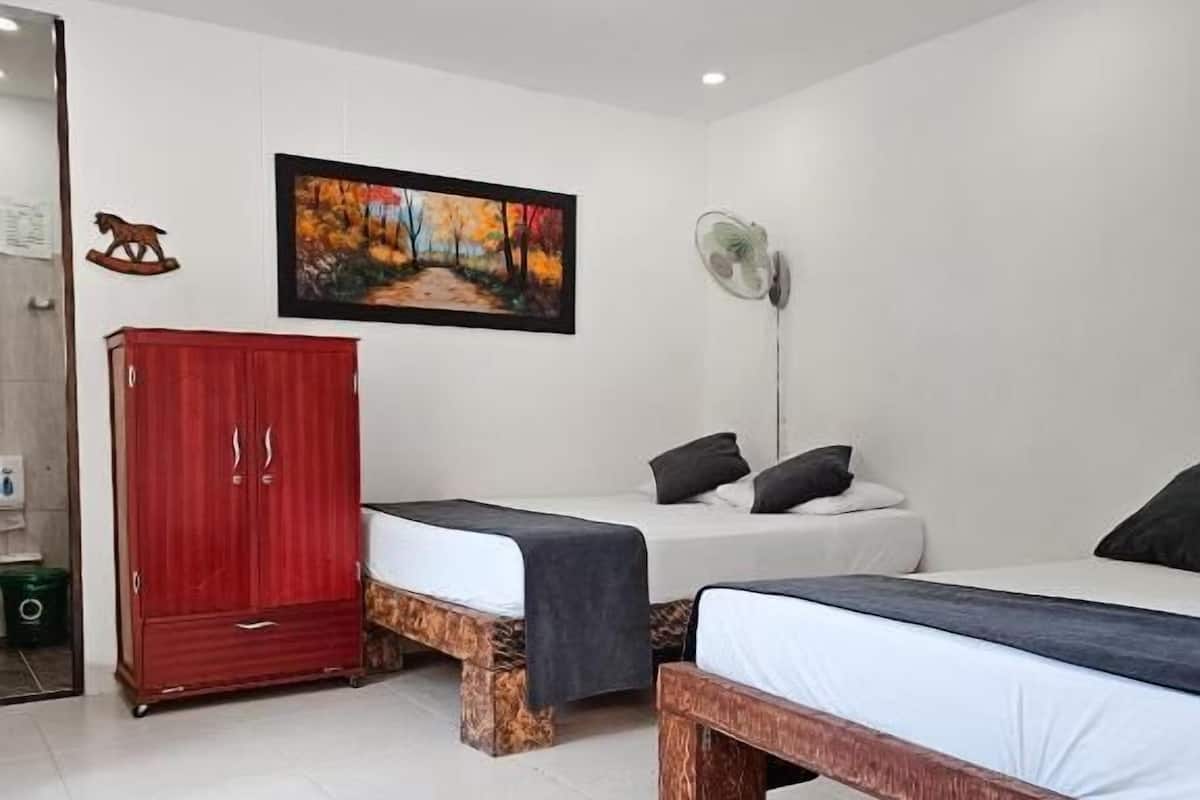 A bedroom with two beds, a red wardrobe, a painting, and a standing fan.