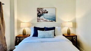 Ein ordentlich bezogenes Bett mit weißer Daunendecke und zwei Kissen, flankiert von zwei Nachttischen mit Lampen, in einem Zimmer mit einem Gemälde von Meereslandschaft an der Wand.