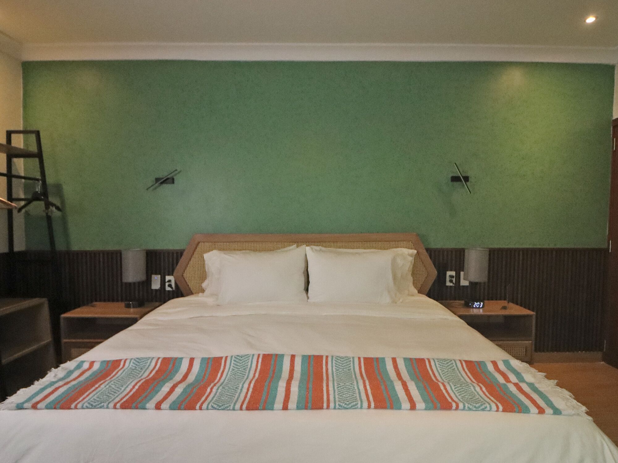 Quarto de hotel com uma cama grande, paredes verdes, duas mesas de cabeceira e um cobertor listrado.