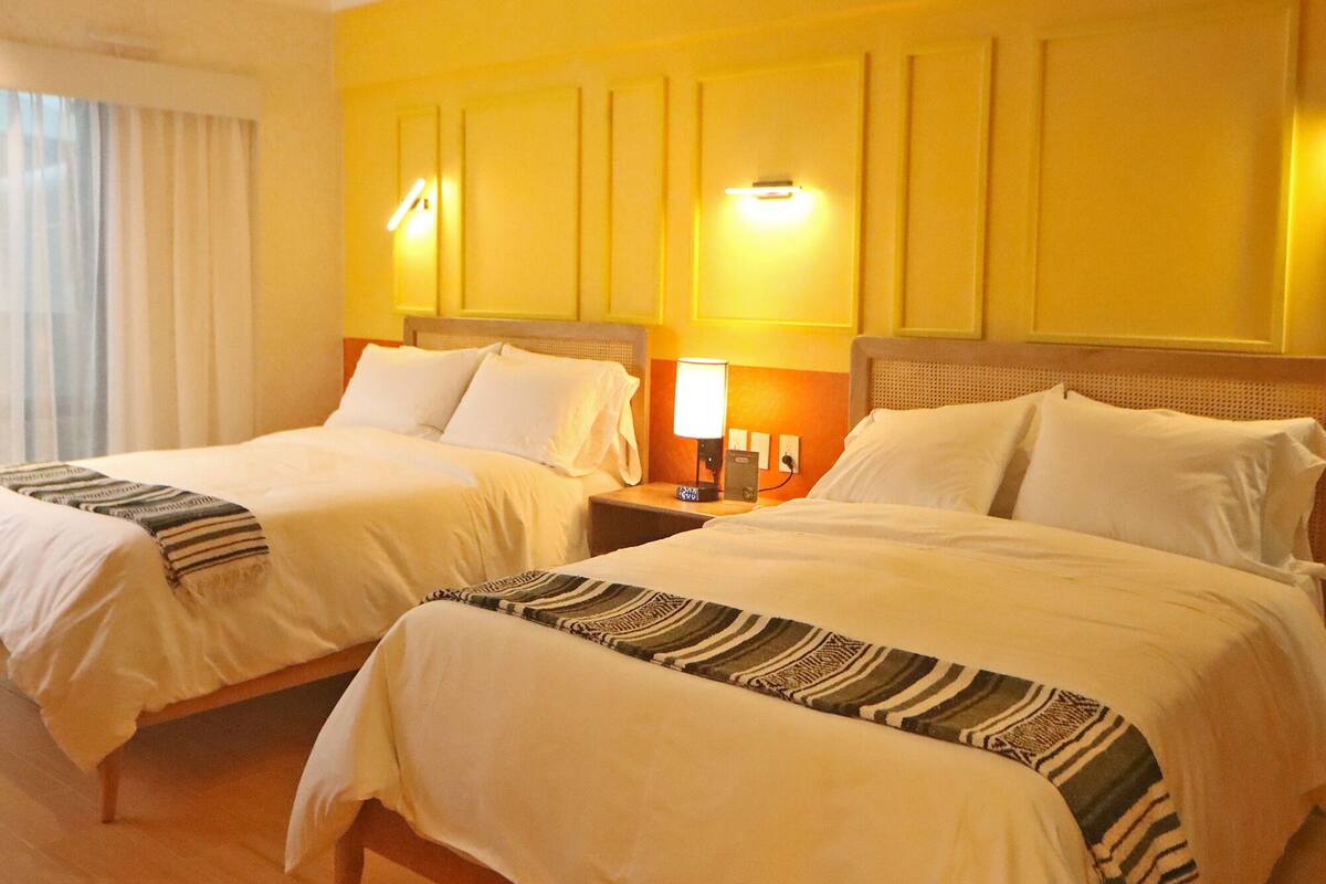 A hotel room with two beds, each with white linens and striped bedspreads. There are bedside lamps and a wall-mounted light fixture.
