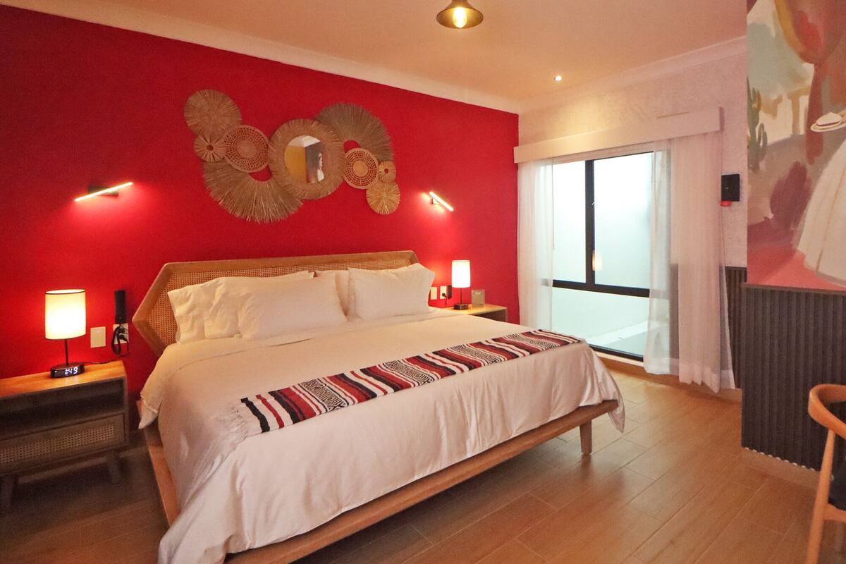 A hotel room with a large bed, two bedside tables with lamps, a wall clock, and a large window with curtains.