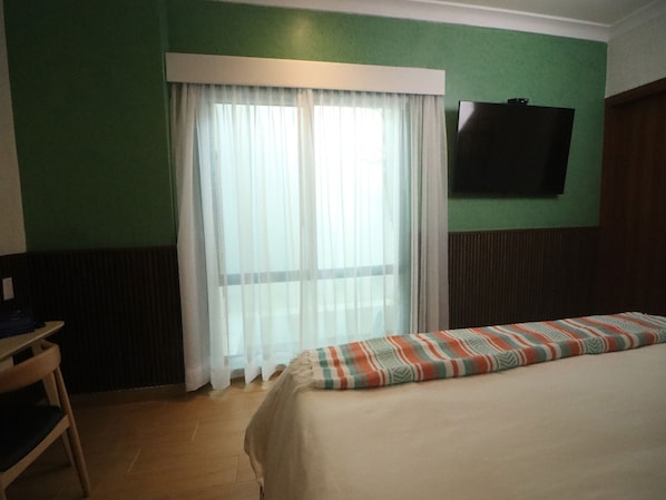 Ett hotellrum med en säng, en tv på väggen och ett fönster med tunna, genomskinliga gardiner.