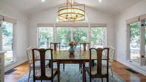 A dining room with a wooden table, six chairs, a chandelier, and large windows with a view of trees.