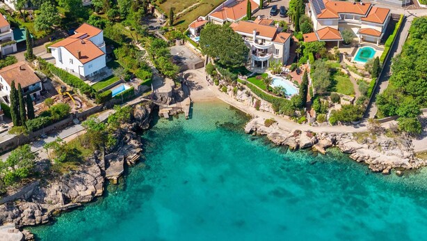 Vue aérienne d’une zone résidentielle côtière, avec des eaux turquoise limpides, des rivages rocheux et des maisons aux toits recouverts de tuiles rouges.