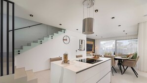 Une cuisine moderne dotée d’un îlot central, d’un coin repas avec une table et des chaises, d’un escalier avec des rampes en verre et d’une grande fenêtre qui laisse entrer la lumière naturelle.