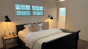Ein ordentlich bezogenes Bett mit weißen Leinen und gemusterten Kissen, ein Nachttisch mit Lampe und ein Fenster mit Blick auf Bäume.