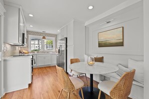 A modern kitchen with white cabinets, a round dining table, wicker chairs, and a large window.