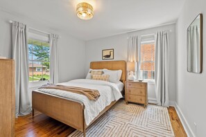 A bedroom with a wooden bed, a nightstand, a lamp, and a view of a brick building through the window.