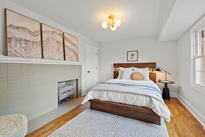 Een slaapkamer met een houten bed, witte muren, een open haard en kunst aan de muur.