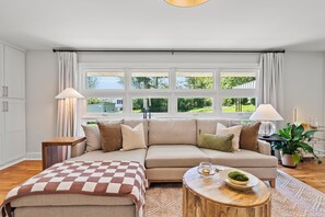 A modern living room with a beige sofa, a wooden coffee table, and a large window with curtains.