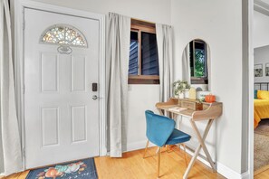 Ein Zimmer mit weißer Tür, einem kleinen Schreibtisch mit Stuhl, einem Fenster mit Vorhängen und einem Bett mit gelber Tagesdecke.