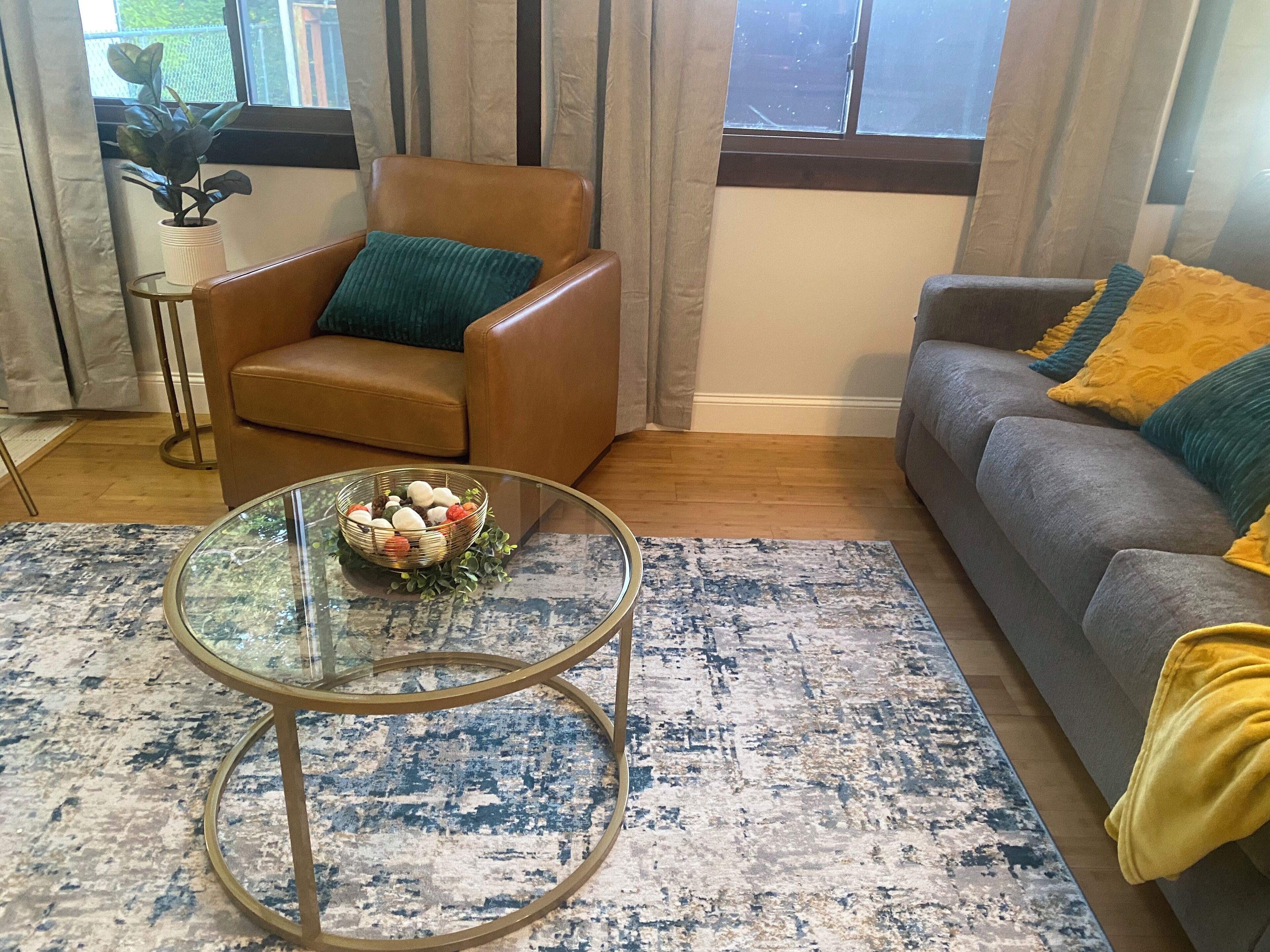 A living room with a brown leather armchair, a grey sofa, a glass coffee table with a woven basket, and a blue and white patterned rug.