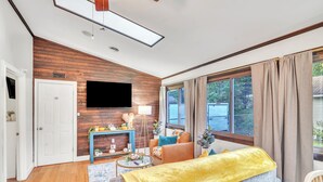 A modern living room with a wooden accent wall, a flat-screen TV, a sofa with a yellow throw, and a large window with curtains.