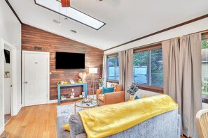 A modern living room with a wooden accent wall, a flat-screen TV, a sofa with a yellow throw, and a large window with curtains.