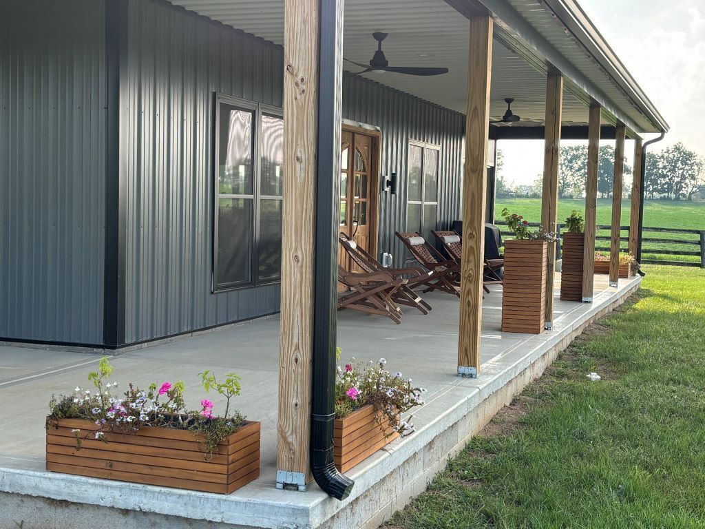 A covered outdoor area with wooden chairs and tables, a wooden planter with flowers, and a view of a green field.