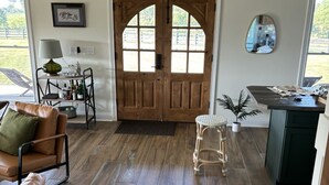 A spacious living room with a large wooden door, a leather sofa, a small stool, and a dining area with a table and chairs.
