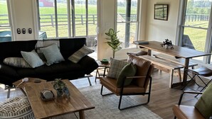 A spacious living room with a black leather sofa, wooden coffee table, and a dining area with a wooden table and chairs. Large windows provide a view of the outdoors.