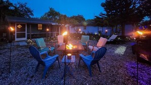 A patio area with a fire pit surrounded by chairs, a covered seating area, and a building in the background. - Robinhood Beach Villa (Sarasota)