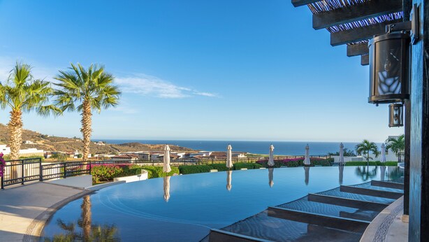 A swimming pool with a view of the ocean, palm trees, and a building with a balcony. - Quivira PH 3BR Premium Condo w/ Golf Cart (Cabo San Lucas)