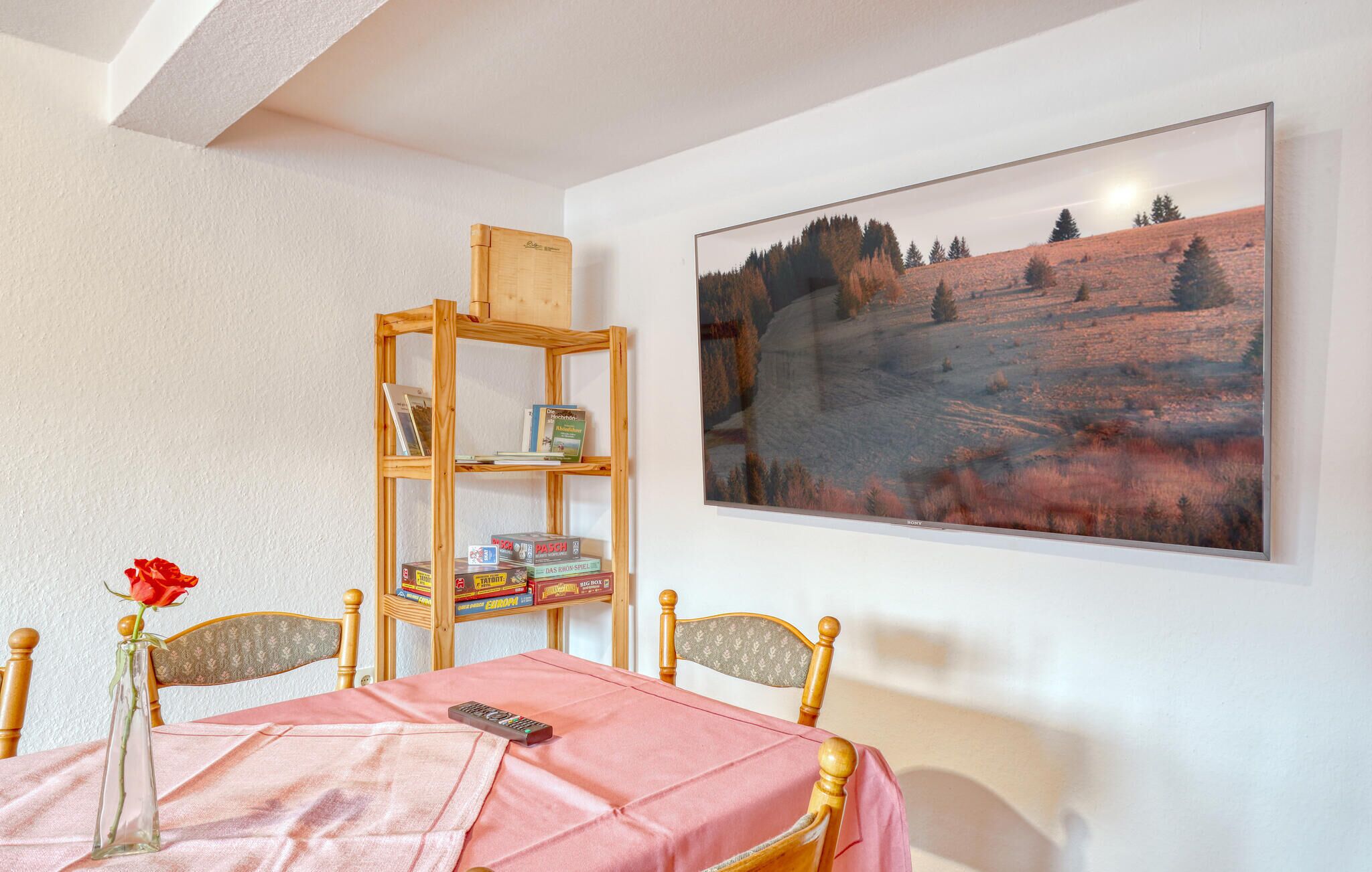 A room with a dining table, chairs, a bookshelf, a vase with a rose, and a framed landscape painting.