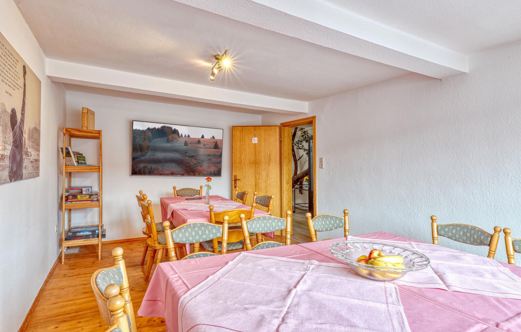 A dining area with a table set for four, wooden chairs, a bookshelf, and a painting on the wall.