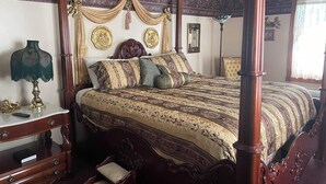 Ein Bett mit Baldachin, reich verziert mit Schnitzereien, ein Nachttisch aus Holz mit einer Lampe und ein Sessel mit gepolstertem Sitz.