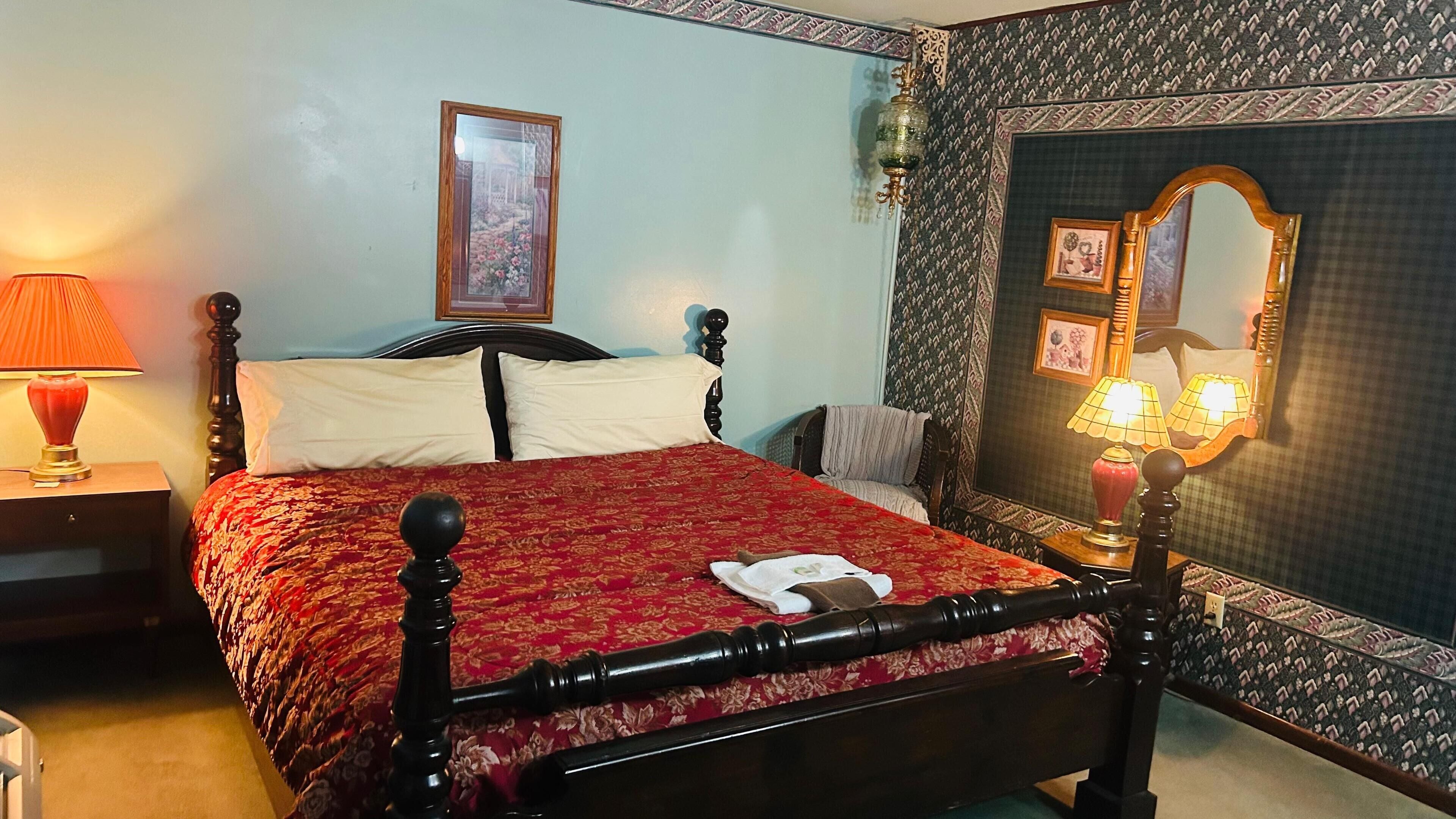 Un lit bien fait, recouvert d’une couverture rouge, avec deux oreillers et une serviette blanche. On y voit une tête de lit en bois, une table de chevet avec une lampe et un miroir accroché au mur.