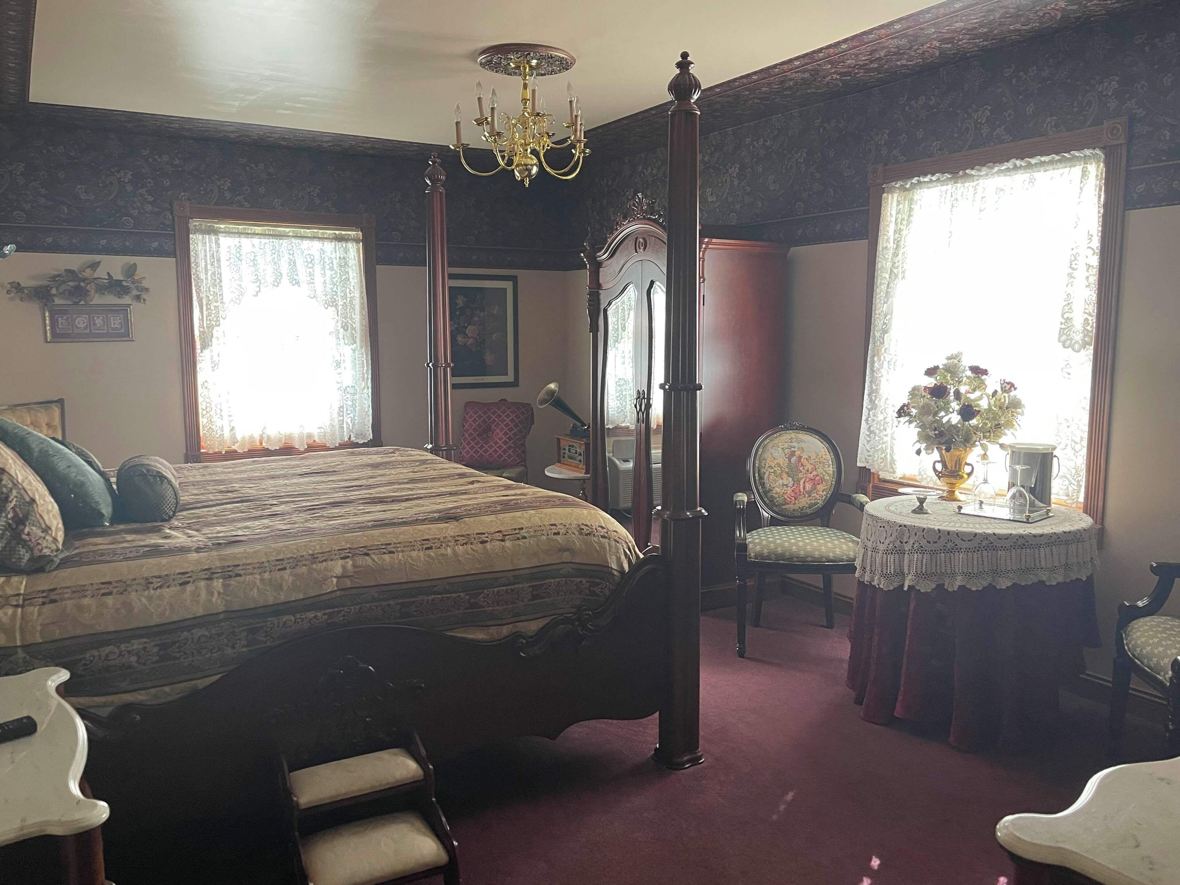 A traditional bedroom with a four-poster bed, a chandelier, a vintage radio, and a floral arrangement by the window.