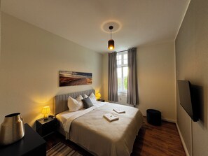 A hotel room with a bed, a nightstand, a lamp, a painting, a window with curtains, and a TV on the wall.