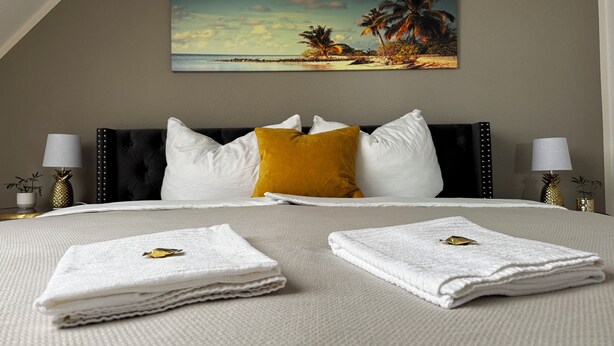 Una camera d'albergo con un letto, cuscini, un quadro raffigurante una spiaggia e due abat-jour.
