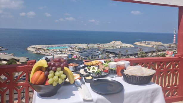 A balcony with a table set for breakfast, overlooking a coastal view with a lighthouse and beach. - Manara Riva Suites  (Beirut)