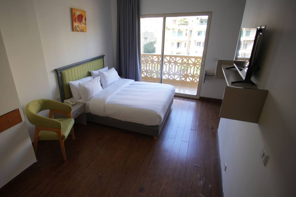 Habitación de hotel con cama, una silla, escritorio con televisor y balcón con vista a edificios.