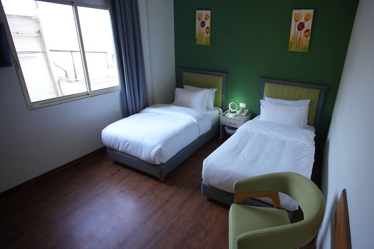 Habitación de hotel con dos camas individuales, una mesita de noche con lámpara, una silla y una ventana con cortinas.