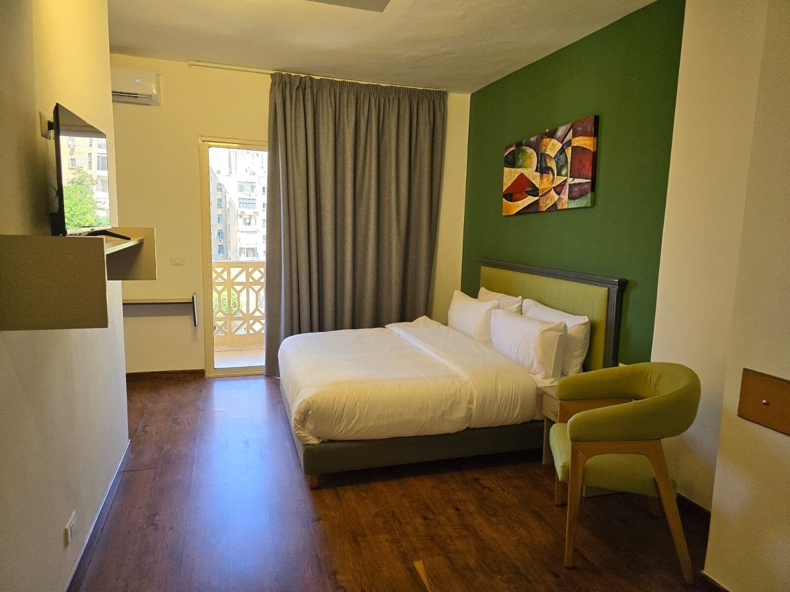 A hotel room with a bed, a green wall, a painting, a chair, and a view of buildings through a window.