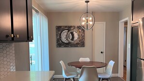 Een moderne keuken met donkere kastjes, een marmeren aanrechtblad, een eettafel met witte stoelen, een kroonluchter en een groot abstract schilderij aan de muur.
