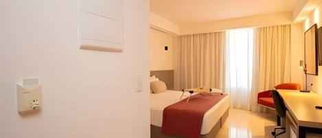 Habitación de hotel con cama, escritorio, silla, ventana con cortinas e interruptor de luz en la pared.