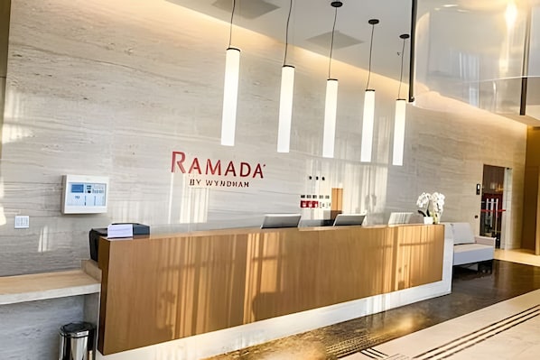 A reception desk with a wooden finish, a wall-mounted sign reading 'RAMADA BY WYNDHAM', and pendant lighting.