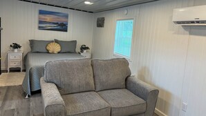 A bedroom with a bed, a grey sofa, a painting on the wall, and a window with blinds.