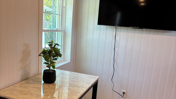 A dining area with a marble table, black chairs, a potted plant, a window with a view of greenery, and a mounted TV.