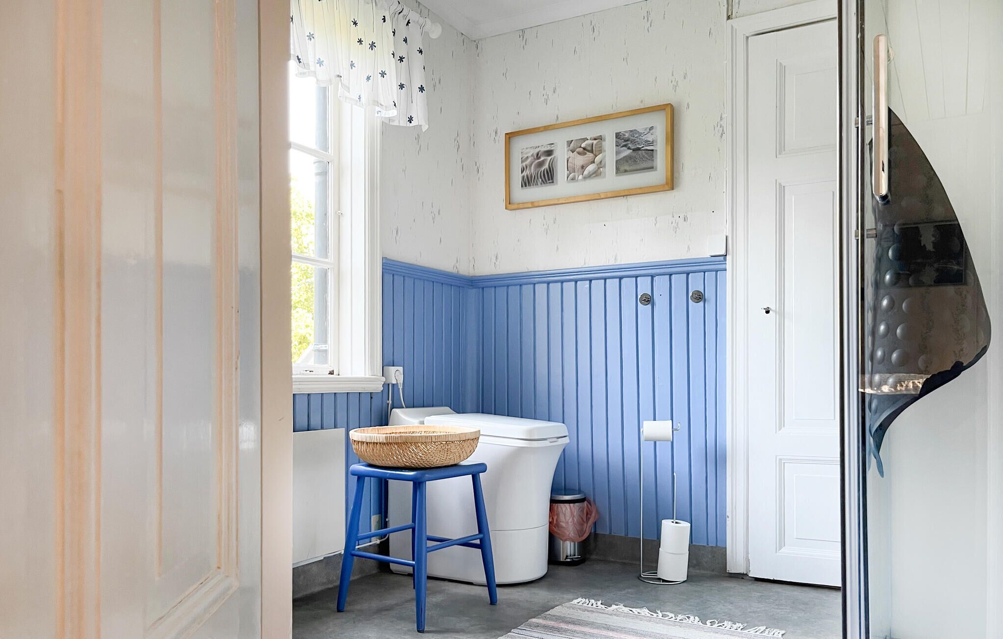 Un baño pequeño con paneles inferiores azules, un inodoro blanco, un taburete azul con una cesta y un cuadro enmarcado en la pared.