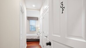 Un pasillo estrecho con una cama al fondo, una puerta con un número y una ventana con persianas.