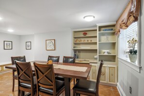A dining area with a wooden table and chairs, a kitchen area with shelves, and a window with a plant. - Spacious 6BR En Suite Rittenhouse Home! Sleeps 14! (Philadelphia)