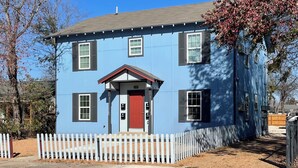 Una casa azul de dos pisos con puerta roja, cerca de piquetes blancos y un pequeño porche.