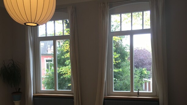 Ein Zimmer mit Holzboden, einem runden Couchtisch, einer Bank, einer Topfpflanze und einem großen Fenster mit Vorhängen.