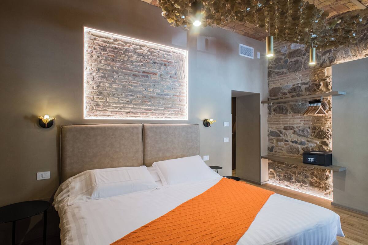 A modern bedroom with a brick wall, a bed with white and orange bedding, and a wall-mounted light fixture.